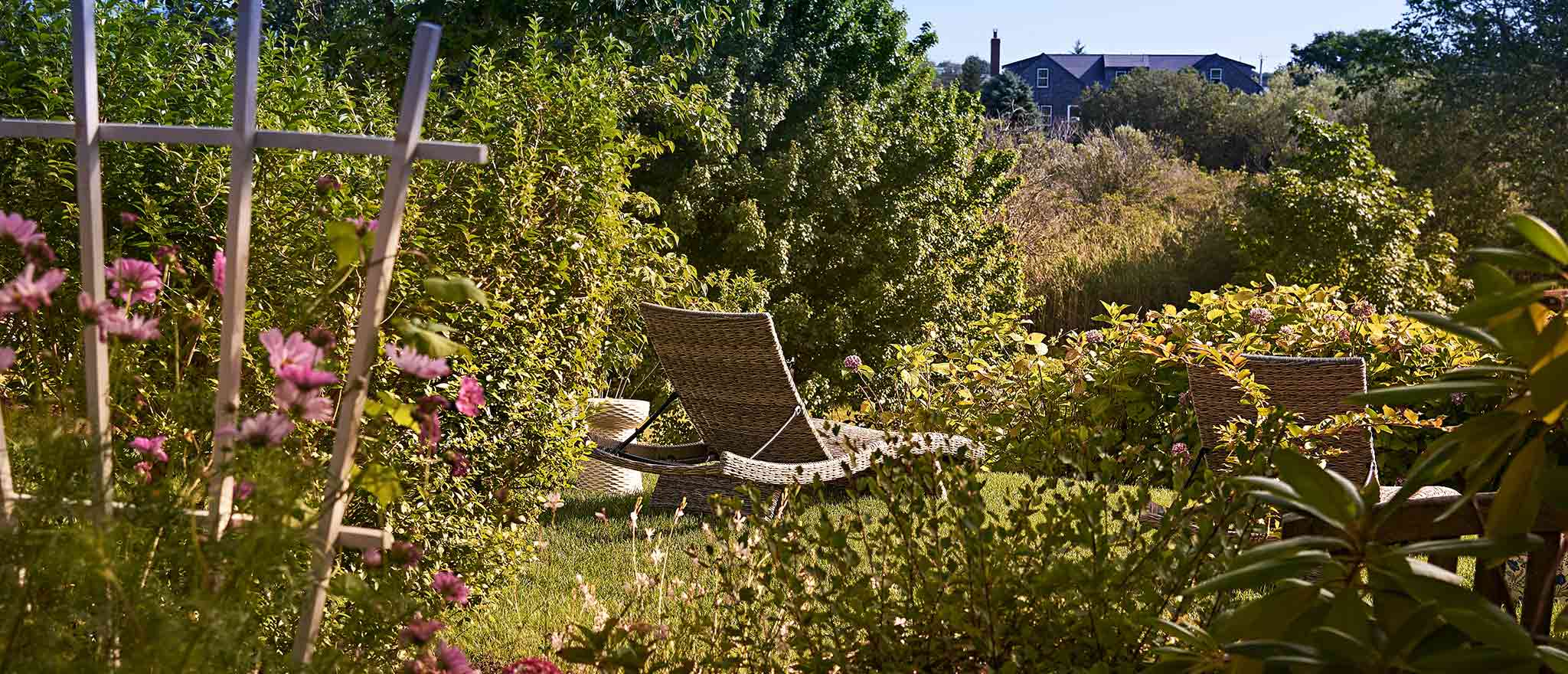 A wicker chaise lounge in the garden surrounded by grass and flowers